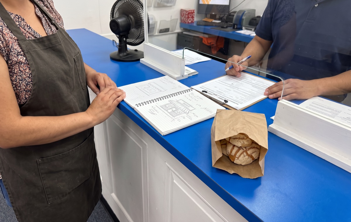 Una señora con delantal muestra su libreta con el dibujo de su panadería mientras le revisan el formulario para abrir el negocio