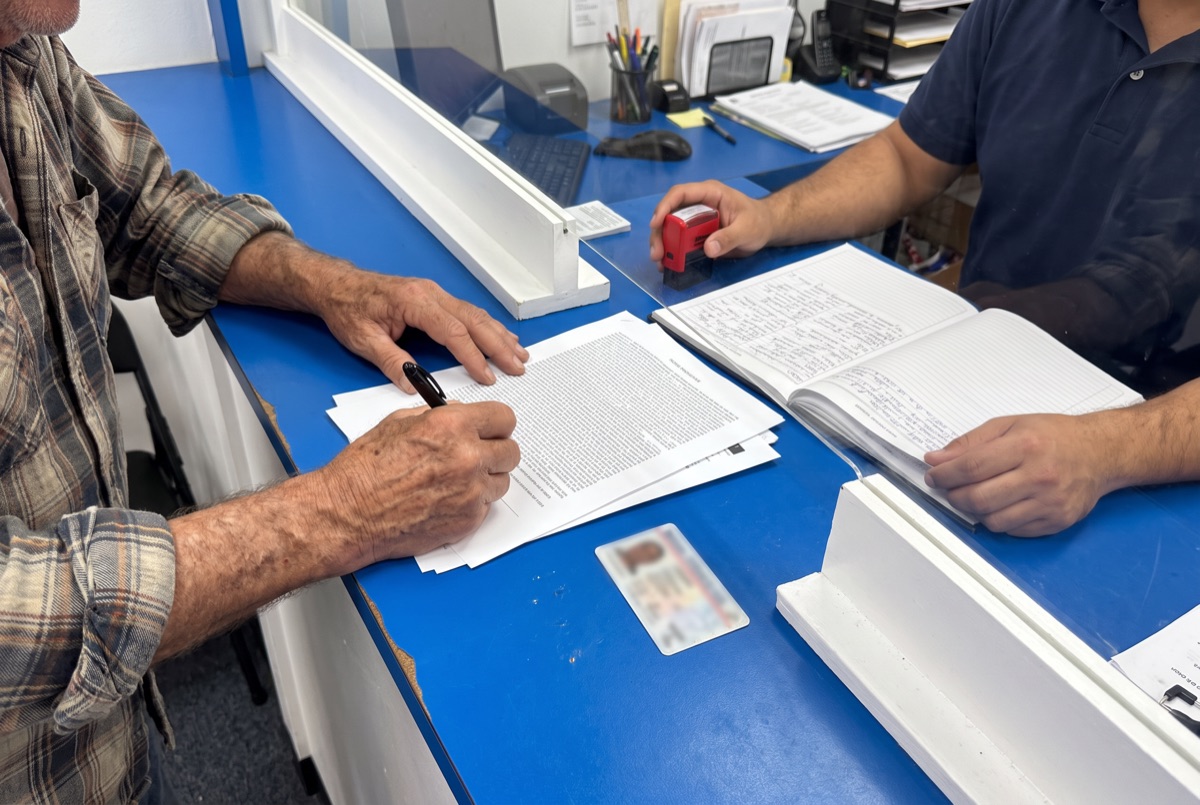 Un señor firma un documento en el mostrador mientras le preparan el sello del notario